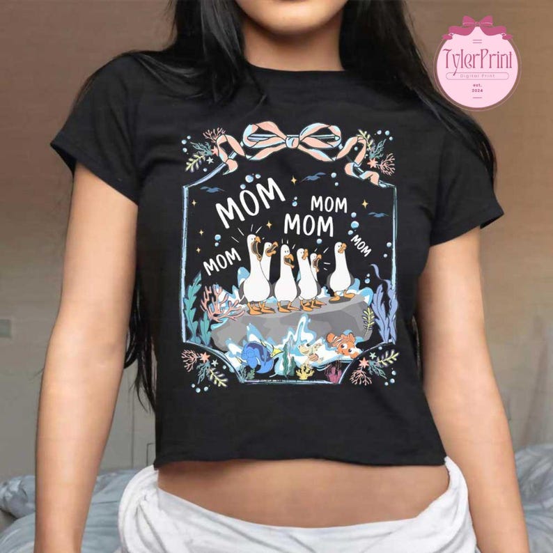 May include: Black crop top with a graphic featuring five white geese with orange beaks and feet, standing on a rock. The word "MOM" is repeated around the geese. A pink bow and floral accents complete the design.