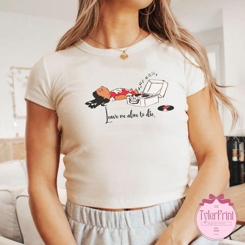 Puede incluir: Camiseta color crema con un gr&aacute;fico de una persona acostada junto a un tocadiscos. La ilustraci&oacute;n incluye el texto "Leave me alone to die". Un disco de vinilo est&aacute; al lado del tocadiscos. La camiseta tiene mangas cortas.