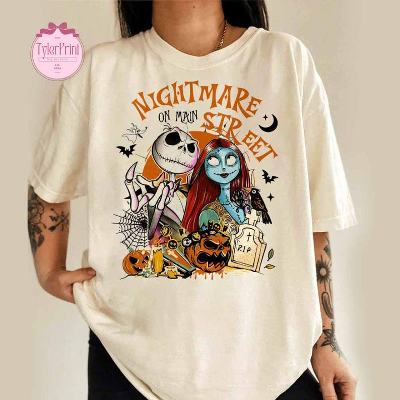 K&ouml;nnte beinhalten: Cremefarbenes T-Shirt mit einer Grafik aus "The Nightmare Before Christmas". Das Design zeigt Jack Skellington, Sally und andere Charaktere mit dem Text "Nightmare on Main Street". Das Design enth&auml;lt auch K&uuml;rbisse, Flederm&auml;use und einen Grabstein.