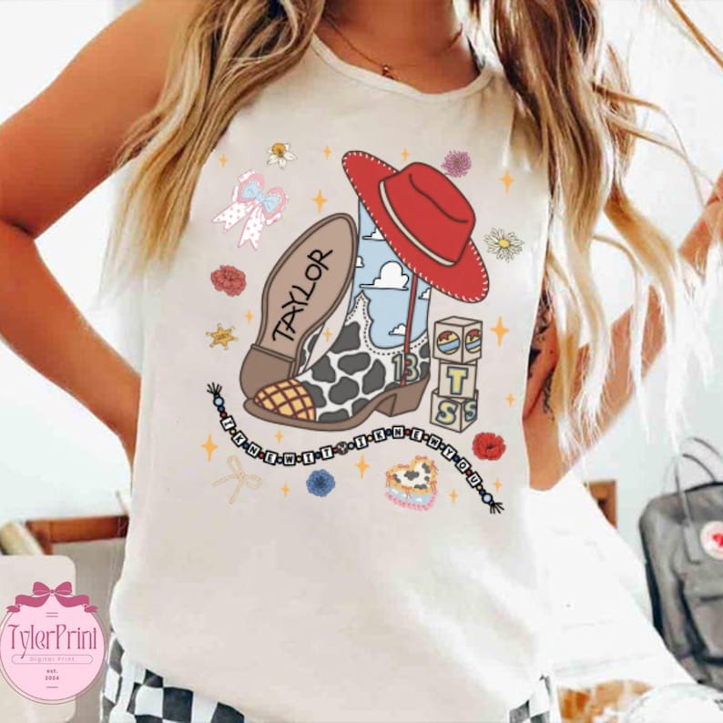 May include: A white sleeveless shirt featuring a graphic print of a red cowboy hat, a cow-print boot with the name Taylor, and stacked blocks with the letters T, S, and the number 13. The design includes a beaded necklace and small floral accents.