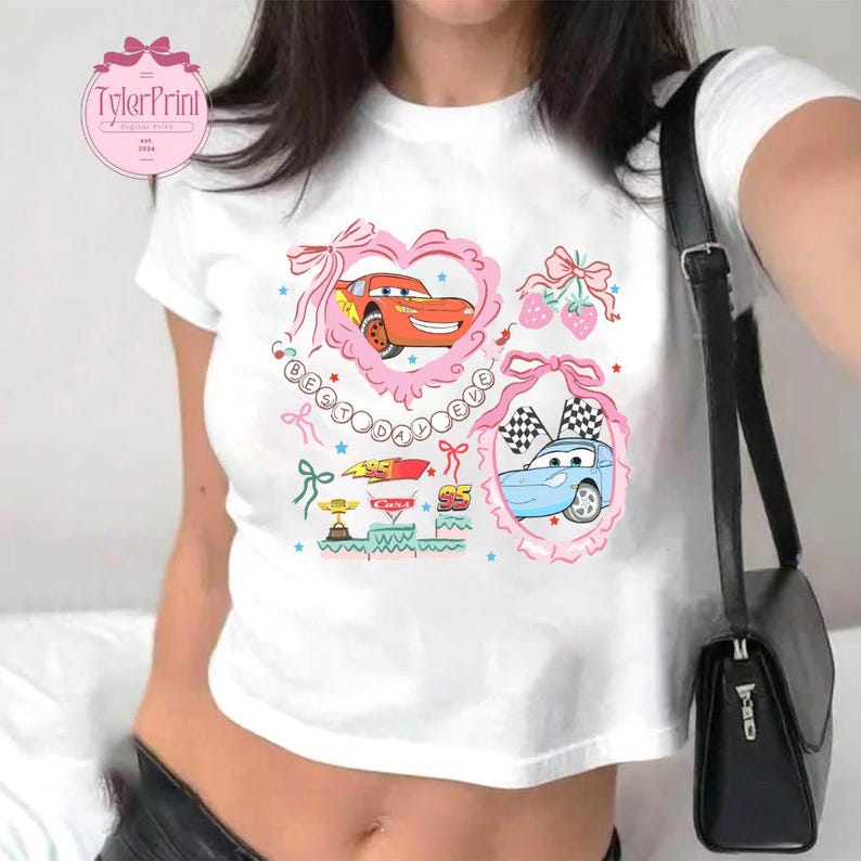 Puede incluir: Top blanco corto con un dise&ntilde;o gr&aacute;fico rosa y azul con coches de dibujos animados, corazones y el texto "Best Day Ever".