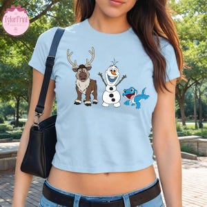 Peut inclure: T-shirt bleu clair avec des personnages de dessins anim&eacute;s du film La Reine des Neiges : un renne, un bonhomme de neige et une cr&eacute;ature bleue ressemblant &agrave; un l&eacute;zard. Le t-shirt a un col rond et des manches courtes. Le logo TylerPrint est visible.