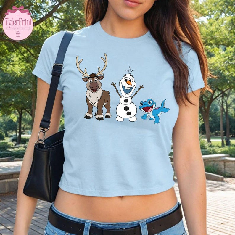 Peut inclure: T-shirt bleu clair avec des personnages de dessins anim&eacute;s du film La Reine des Neiges : un renne, un bonhomme de neige et une cr&eacute;ature bleue ressemblant &agrave; un l&eacute;zard. Le t-shirt a un col rond et des manches courtes. Le logo TylerPrint est visible.