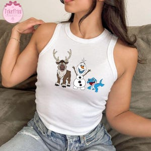 Peut inclure: D&eacute;bardeur blanc avec des illustrations de dessins anim&eacute;s d'un renne, d'un bonhomme de neige et d'un l&eacute;zard bleu. Le renne a des bois et une fourrure brune. Le bonhomme de neige a un nez en carotte et des bras en b&acirc;ton.