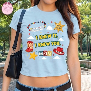 May include: A light blue t-shirt featuring a graphic print with a beaded bracelet, a yellow sheriff star, red cowboy boots, a red cowboy hat, and alphabet blocks spelling TS5. The text reads I KNEW IT, I KNEW YOU, INFINITY AND BEYOND.