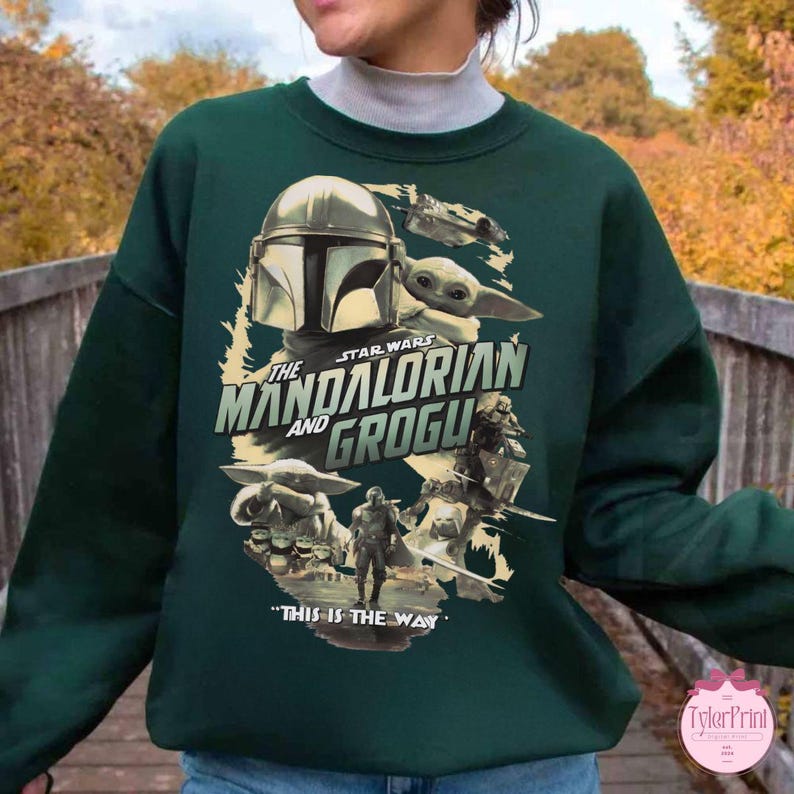 May include: Dark green sweatshirt featuring a graphic design from the Star Wars series, "The Mandalorian and Grogu." The design includes images of the Mandalorian, Grogu, and the text "This is the Way."