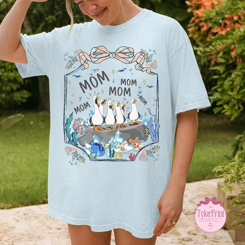 May include: Light blue t-shirt featuring a whimsical design with the word "MOM" repeated around a framed illustration. The illustration depicts a group of cartoon ducks standing on a rock, surrounded by aquatic elements.