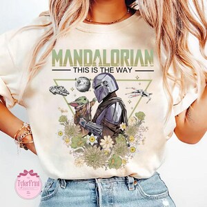 May include: Cream-colored t-shirt featuring a graphic of The Mandalorian and Grogu surrounded by flowers. The text "MANDALORIAN - THIS IS THE WAY" is printed above the characters. Includes images of a spaceship and the Death Star.