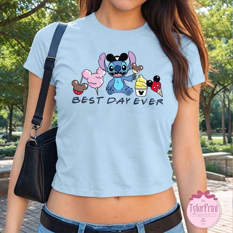 Pode incluir: Uma camiseta cropped azul claro com um personagem de desenho animado do Stitch e o texto "Best Day Ever". O design apresenta o Stitch segurando um cone de sorvete do Mickey Mouse e cercado por outras guloseimas com tema Disney.