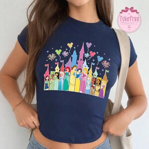 May include: A navy blue cropped t-shirt featuring a colourful graphic of Disney princesses in front of a castle. The graphic includes fireworks, balloons, and the text "TylerPrint".