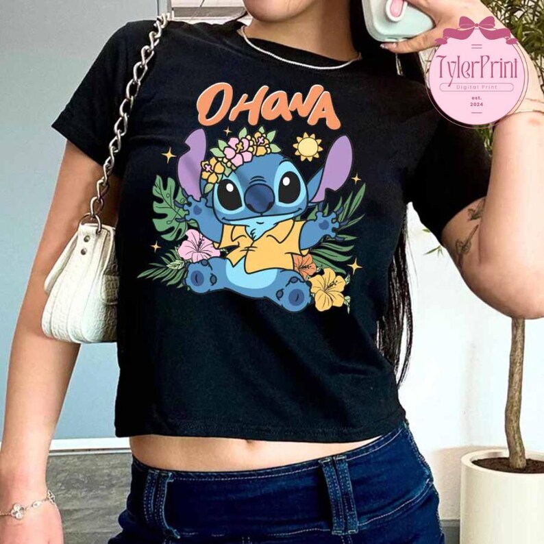 May include: Black crop top featuring a colorful graphic of the character Stitch from Lilo & Stitch, with the word "OHANA" above. Stitch is surrounded by tropical flowers and foliage. The shirt is paired with blue jeans.