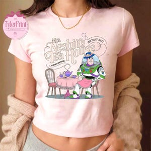 Może przedstawiać: R&oacute;żowa koszulka z kresk&oacute;wkowym wizerunkiem Buzz Lightyeara z Toy Story siedzącego przy stole z czajnikiem i filiżankami do herbaty. Tekst "Mrs. Nesbitt's Tea House" znajduje się nad obrazkiem.