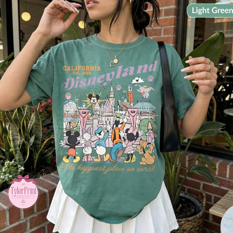 Puede incluir: Camiseta verde claro con un dise&ntilde;o de Disneyland que presenta a Mickey Mouse y sus amigos. La camiseta tiene el texto "California Disneyland Est. 1955" y "The happiest place on earth."