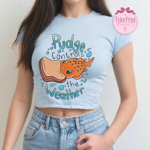 May include: Pale blue crop top featuring the text "Pudge controls the Weather" and a cartoon illustration of an orange fish with blue spots holding a piece of bread. The shirt has short sleeves.