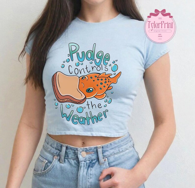 May include: Pale blue crop top featuring the text "Pudge controls the Weather" and a cartoon illustration of an orange fish with blue spots holding a piece of bread. The shirt has short sleeves.