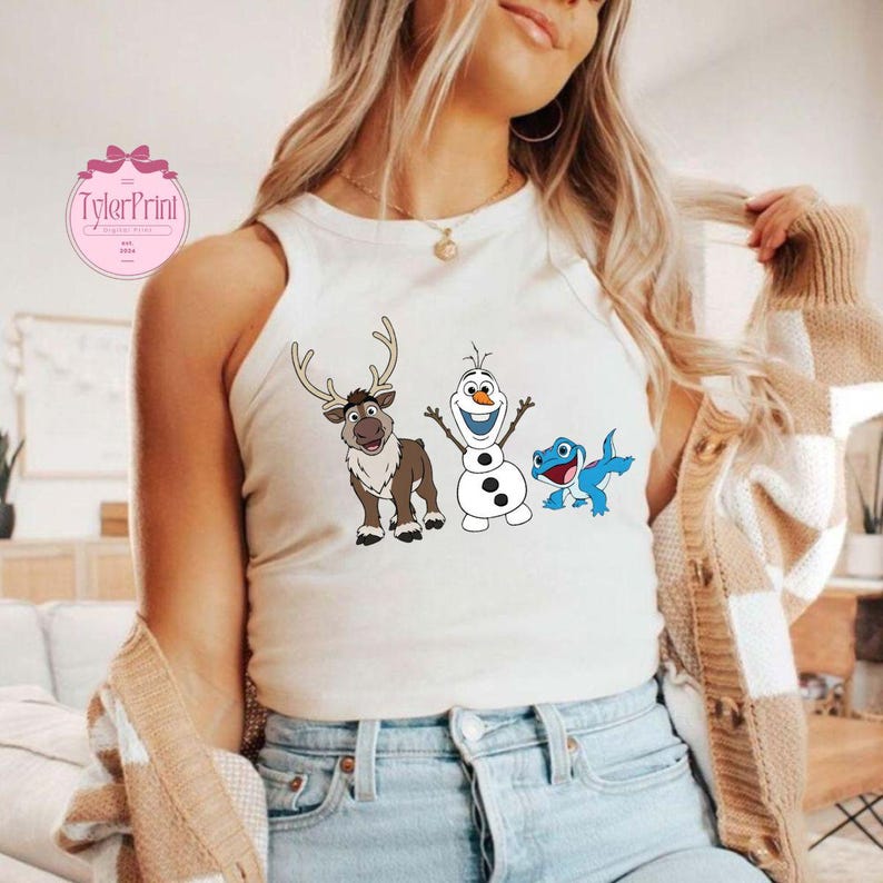 Peut inclure: Un d&eacute;bardeur blanc avec un graphisme color&eacute; de personnages de dessins anim&eacute;s. Le graphisme comprend un renne, un bonhomme de neige et un l&eacute;zard bleu. Le d&eacute;bardeur est sans manches et en mati&egrave;re souple. Le texte "TylerPrint" est visible en haut &agrave; gauche.