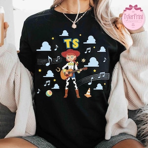 May include: A black t-shirt featuring a graphic of a cartoon cowgirl playing an acoustic guitar. The design includes the text TS, blue clouds, yellow stars, musical notes, a horseshoe, a colorful ball, and an orange traffic cone on the front.