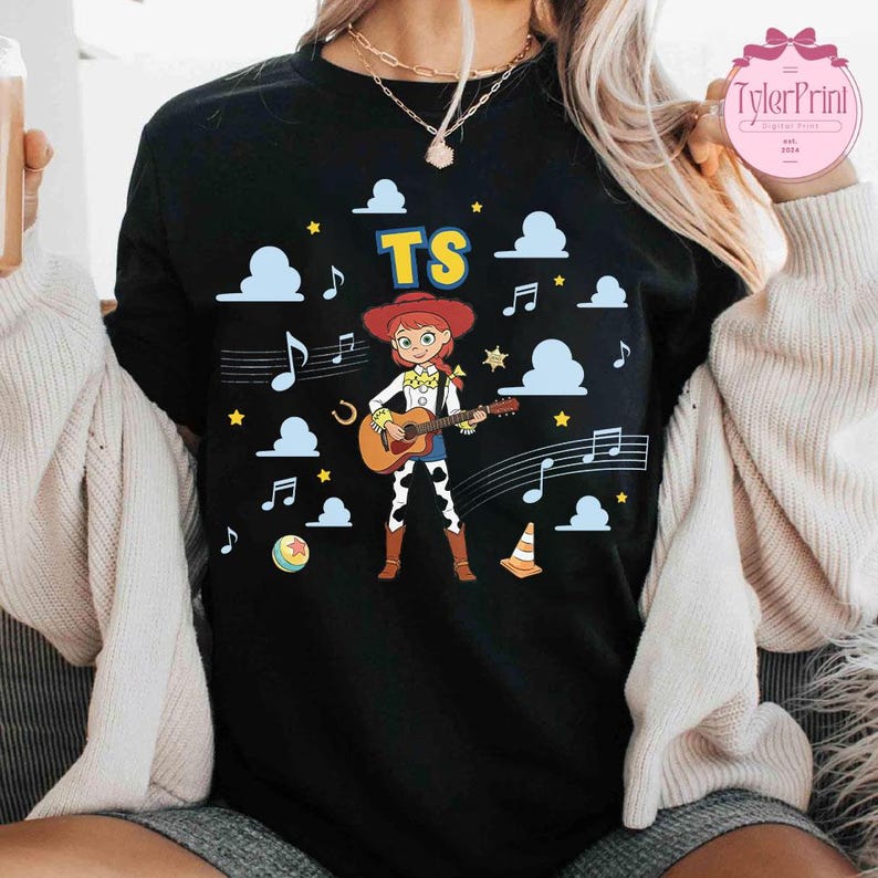 May include: A black t-shirt featuring a graphic of a cartoon cowgirl playing an acoustic guitar. The design includes the text TS, blue clouds, yellow stars, musical notes, a horseshoe, a colorful ball, and an orange traffic cone on the front.