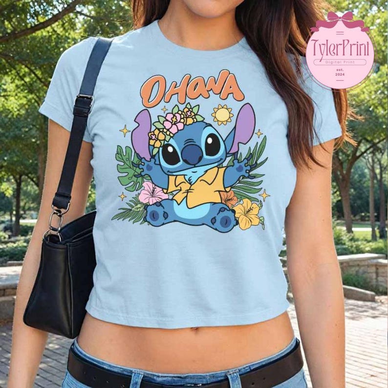 May include: Light blue crop top featuring a colorful graphic of Stitch from Lilo & Stitch, surrounded by tropical flowers and foliage. The word "Ohana" is written above the character. The shirt has short sleeves and a round neckline.