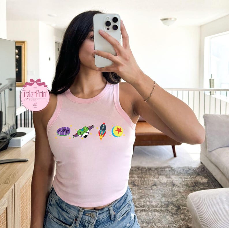 May include: Pink tank top with a graphic print of characters from the movie Toy Story. The characters include Woody, Buzz Lightyear, and the alien. The tank top is cropped and has a scoop neck.