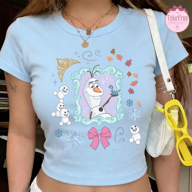 May include: Light blue crop top featuring a cartoon image of Olaf from Frozen. The image includes snowflakes, leaves, and a pink bow. The text "TylerPrint" is visible on the upper right corner. The shirt has short sleeves and a round neckline.