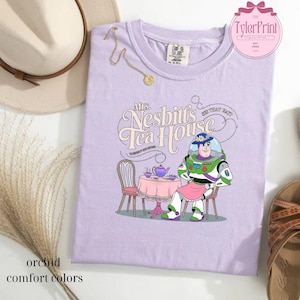 Może przedstawiać: Fioletowy T-shirt z kresk&oacute;wkowym wizerunkiem Buzz Lightyeara z Toy Story siedzącego przy stole z czajnikiem i filiżankami do herbaty. Tekst "Mrs. Nesbitt's Tea House" znajduje się nad obrazkiem.