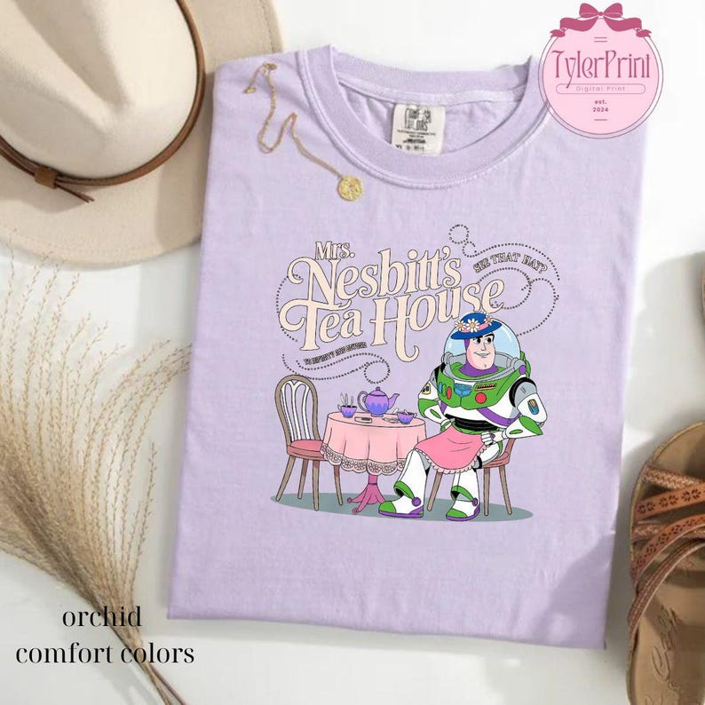 Może przedstawiać: Fioletowy T-shirt z kresk&oacute;wkowym wizerunkiem Buzz Lightyeara z Toy Story siedzącego przy stole z czajnikiem i filiżankami do herbaty. Tekst "Mrs. Nesbitt's Tea House" znajduje się nad obrazkiem.