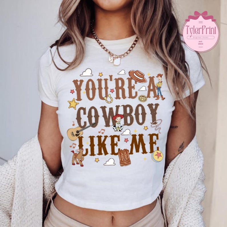 May include: A white short-sleeved t-shirt featuring the brown text "YOU'RE A COWBOY LIKE ME" surrounded by colorful illustrations of a cowboy hat, a guitar, a horse, a boot, stars, clouds, and cartoon characters in a western-themed graphic design.