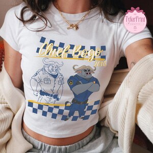 May include: White crop top with a cartoon illustration of a buffalo in a police uniform. The text "Chief Bogo 2016" is printed above the buffalo. The design includes a blue and yellow checkered pattern.