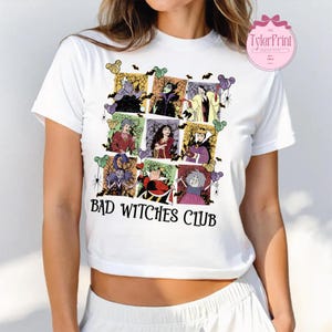May include: White t-shirt featuring a colorful graphic of various cartoon villains in a grid pattern, with bats and spiders. The text "BAD WITCHES CLUB" is printed in black below the graphic.