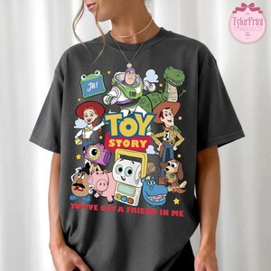 May include: Dark gray t-shirt featuring a colorful graphic of Toy Story characters, including Buzz Lightyear, Woody, and Rex. The words "TOY STORY" are prominently displayed, with the phrase "YOU'VE GOT A FRIEND IN ME" below the characters.