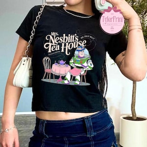 Może przedstawiać: Czarny, obcięty T-shirt z grafiką przedstawiającą kresk&oacute;wkowego kosmicznego strażnika siedzącego przy stole z czajnikiem i filiżankami do herbaty. Tekst na koszulce brzmi "Mrs. Nesbitt's Tea House" i "See that hat?"