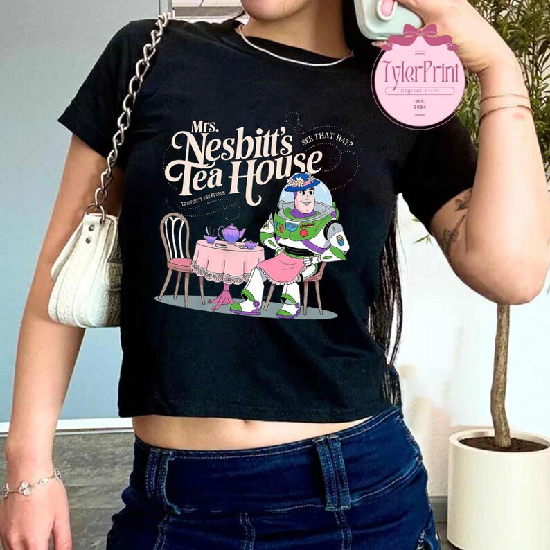 Może przedstawiać: Czarny, obcięty T-shirt z grafiką przedstawiającą kresk&oacute;wkowego kosmicznego strażnika siedzącego przy stole z czajnikiem i filiżankami do herbaty. Tekst na koszulce brzmi "Mrs. Nesbitt's Tea House" i "See that hat?"