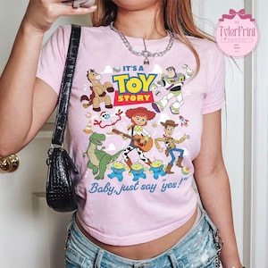 May include: A light pink short-sleeve t-shirt featuring a colorful graphic print of Toy Story characters including Woody, Buzz Lightyear, Jessie, Rex, Bullseye, Forky, and the Aliens. The text reads IT'S A TOY STORY and Baby, just say yes!