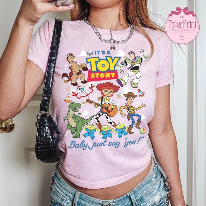 May include: A light pink short-sleeve t-shirt featuring a colorful graphic print of Toy Story characters including Woody, Buzz Lightyear, Jessie, Rex, Bullseye, Forky, and the Aliens. The text reads IT'S A TOY STORY and Baby, just say yes!