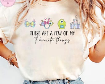 May include: Cream-colored t-shirt featuring the phrase "These Are A Few Of My Favorite Things" in black script. The shirt has colorful cartoon graphics of a bow, a headband, a monster, and a castle. The shirt is a casual, everyday wear item.