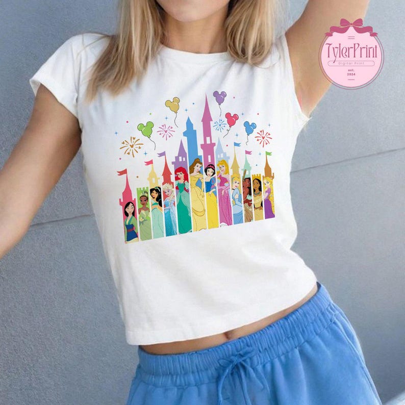 May include: White t-shirt featuring a colourful graphic of Disney princesses in front of Cinderella's Castle. The graphic includes fireworks and balloons.