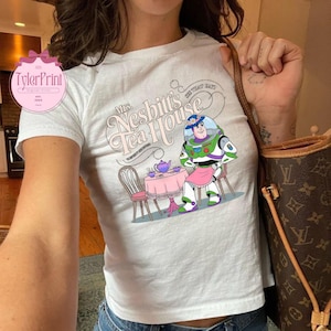 Może przedstawiać: Biała koszulka z nadrukiem przedstawiającym Buzz Lightyeara z Toy Story siedzącego przy stole z czajnikiem i filiżankami do herbaty. Na koszulce widnieje napis "Mrs. Nesbit's Tea House".