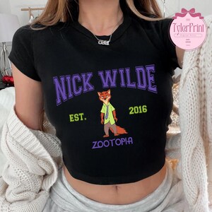 May include: Black crop top featuring the text "NICK WILDE" in purple arched lettering, with a cartoon fox character illustration. Below the text are the words "EST. 2016" and "ZOOTOPIA" in green and purple.