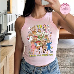 May include: A light pink sleeveless tank top featuring a colorful graphic print of various animated characters in different outfits, the text TS, and the phrase I KNEW IT, I KNEW YOU printed in yellow letters at the bottom of the design.