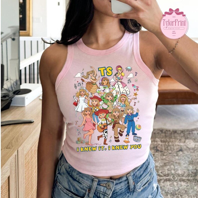 May include: A light pink sleeveless tank top featuring a colorful graphic print of various animated characters in different outfits, the text TS, and the phrase I KNEW IT, I KNEW YOU printed in yellow letters at the bottom of the design.