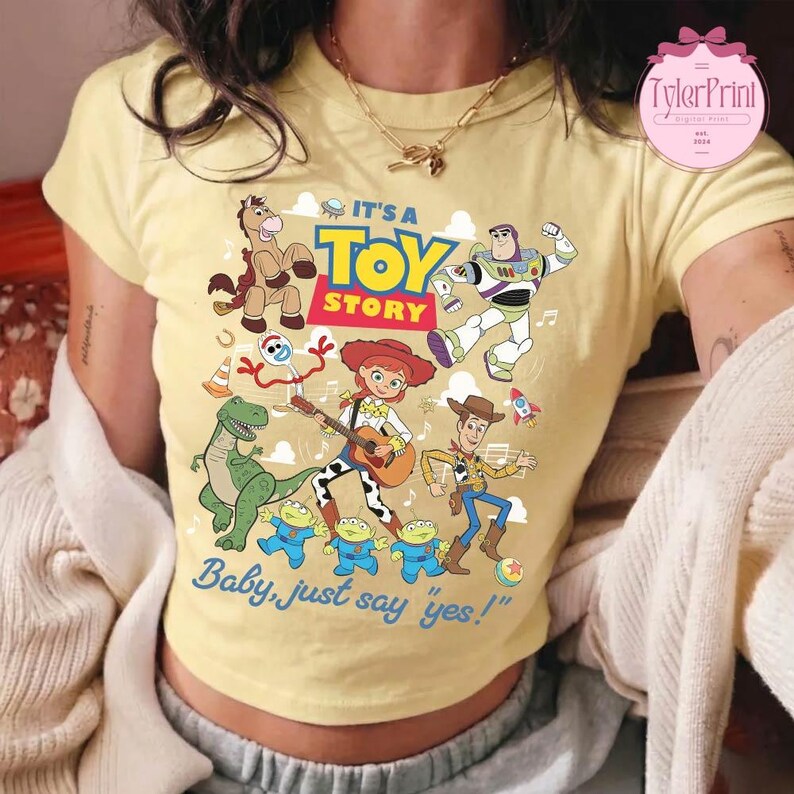 May include: A pale yellow short-sleeved t-shirt featuring a colorful graphic print of Toy Story characters including Woody, Jessie, Buzz Lightyear, Rex, Bullseye, Forky, and three aliens. The text reads IT'S A TOY STORY and Baby, just say yes!