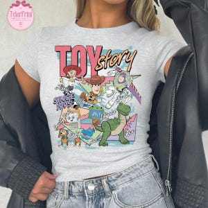 K&ouml;nnte beinhalten: Ein hellgraues Kurzarm-T-Shirt mit einem farbenfrohen Grafikdruck der Toy Story-Figuren wie Jessie, Woody, Buzz Lightyear, Slinky Dog, Rex und anderen, mit dem aufgedruckten Text "TOY story" und "SINCE 1995" auf der Vorderseite.