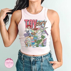 K&ouml;nnte beinhalten: Ein hellrosa geripptes Tanktop mit einem Grafikdruck von Toy Story-Figuren, darunter Woody, Buzz Lightyear, Jessie, Rex, Slinky Dog und andere. Der Text lautet Toy Story, Since 1995, Jessie, Woody, Buzz, Slinky, Smarty Pants, Lilypad und Rex.
