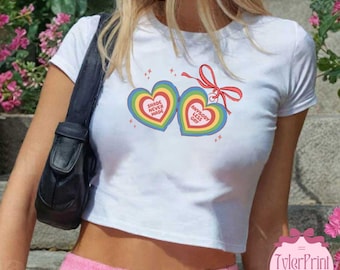 May include: White crop top featuring two rainbow heart graphics with the phrases "SHADE NEVER MADE" and "ANYBODY LESS GAY". A red ribbon connects the hearts. The shirt is a casual style.