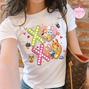 May include: White t-shirt featuring a colorful graphic design with the text "XO" in green and pink, decorated with polka dots. The design includes cartoon characters in heart-shaped frames and coffee cups.