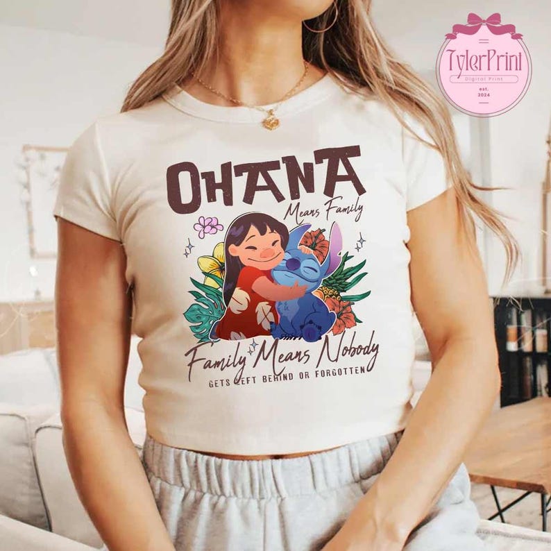 May include: Cream-colored t-shirt featuring a graphic of Lilo and Stitch hugging, with the words "Ohana Means Family" and "Family Means Nobody Gets Left Behind or Forgotten". The design includes flowers and tropical leaves.