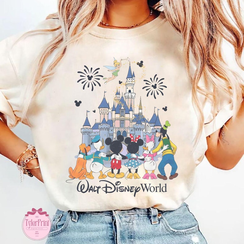 Puede incluir: Camiseta color crema con una colorida ilustraci&oacute;n de personajes de Disney frente a un castillo. El dise&ntilde;o incluye Mickey Mouse, Minnie Mouse, Donald Duck, Daisy Duck, Goofy y Campanilla. Las palabras "Walt Disney World" est&aacute;n impresas debajo.