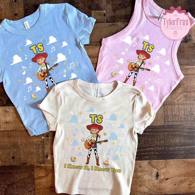 May include: Three shirts in light blue, light pink, and cream featuring a graphic of a cowgirl character playing a guitar. The design includes the text TS, musical notes, clouds, and small balls. The cream shirt also includes the text I Knew It, I Knew You.