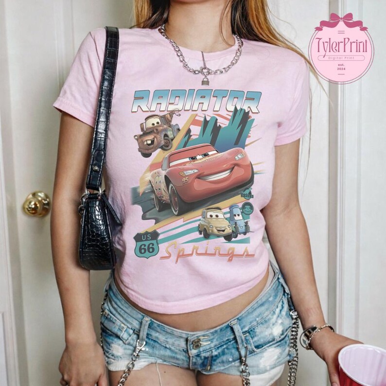 May include: A light pink short-sleeve t-shirt featuring a graphic print of characters from the movie Cars, including Lightning McQueen and Mater. The design includes the text Radiator Springs and a US 66 road sign graphic on the front center.
